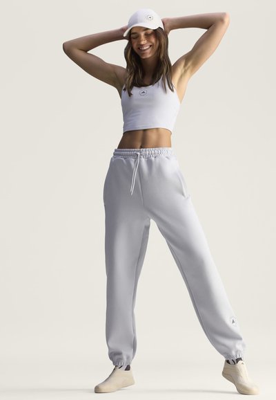 Νέα γυναίκα που φοράει ολοκληρωμένο λευκό αθλητικό ρουχισμό, στέκεται με τα χέρια πίσω από το κεφάλι και χαμογελάει σε απλό φόντο.