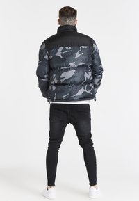 Giacca a vento con motivo camouflage in nero e grigio, con una parte superiore nera uniforme, caratterizzata da un colletto alto, una texture trapuntata e chiusura con zip.