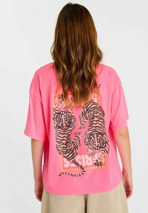 Persoon met lang bruin haar die een losse roze T-shirt draagt met twee zwarte en oranje tijgers en lichtbeige broek.