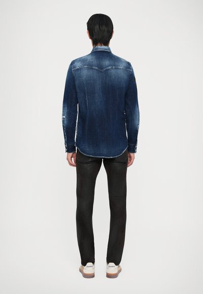 Mann mit dem Rücken zugewandt, trägt ein ausgewaschenes blaues Denim-Hemd, schwarze Slim-Fit-Hosen und weiße Sneakers mit beigen Sohlen vor einem schlichten weißen Hintergrund.