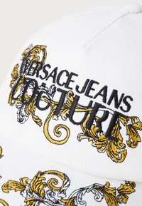 Cappellino bianco in cotone con ricamo nero e oro del testo "Versace Jeans Couture" e ornati disegni floreali. Design classico con visiera curva.