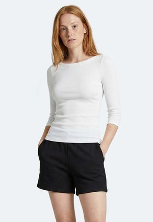 Rothaarige Frau, die ein eng anliegendes weißes Langarmshirt und lockere schwarze Shorts trägt, steht mit den Händen in den Taschen vor einem schlichten Hintergrund.