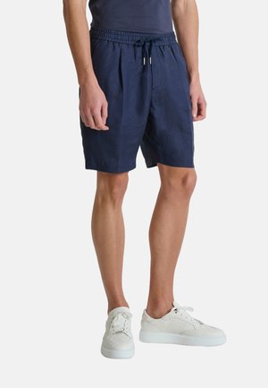 Hombre que lleva puestos unos pantalones cortos azul marino con cordón y bolsillos laterales, y zapatillas blancas, de pie frente a un fondo sencillo.