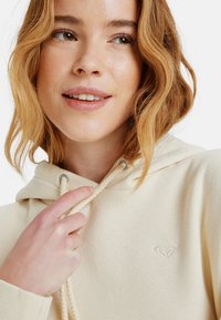 Crème-kleurige hoodie in zachte stof, met een klein geborduurd logo op de borst en gevlochten koordjes met metalen accenten.
