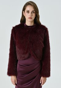 Giacca cropped in pelliccia sintetica bordeaux con maniche lunghe, caratterizzata da una texture morbida e un colletto rotondo, abbinata a un vestito in satin dello stesso colore.