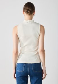 Ärmelloses Top mit hohem Kragen aus cremefarbenem Rippmaterial. Glatte Textur, taillierte Passform. Im Alltag mit blauen Jeans getragen, Rückansicht sichtbar.