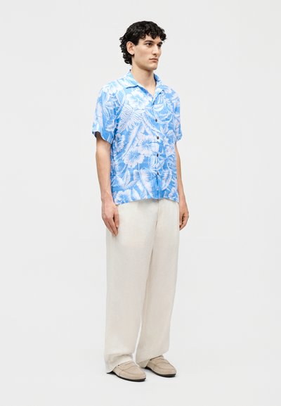 Fiatal férfi áll, kék-fehér virágmintás rövid ujjú inget, bő fehér nadrágot és bézs slip-on cipőt visel, egyszínű háttér előtt.