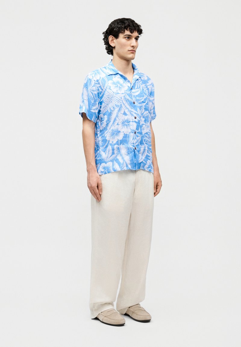 Jonge man staand, gekleed in een blauw-wit bloesje met korte mouwen met bloemenpatroon, losse witte broek en beige instapschoenen tegen een effen achtergrond.