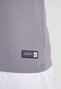 Šedé atletické tričko s krátkým rukávem, na kterém je našitá emblémová nálepka s nápisem "AUTENTICKÝ FOOTBALL" a logo Nike. Bílé síťované akcenty.