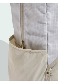 Mochila beige con tela gris claro texturizada, patrón de cuadrícula y un bolsillo lateral. Cuenta con cierre de cremallera y detalles de tela en el diseño.