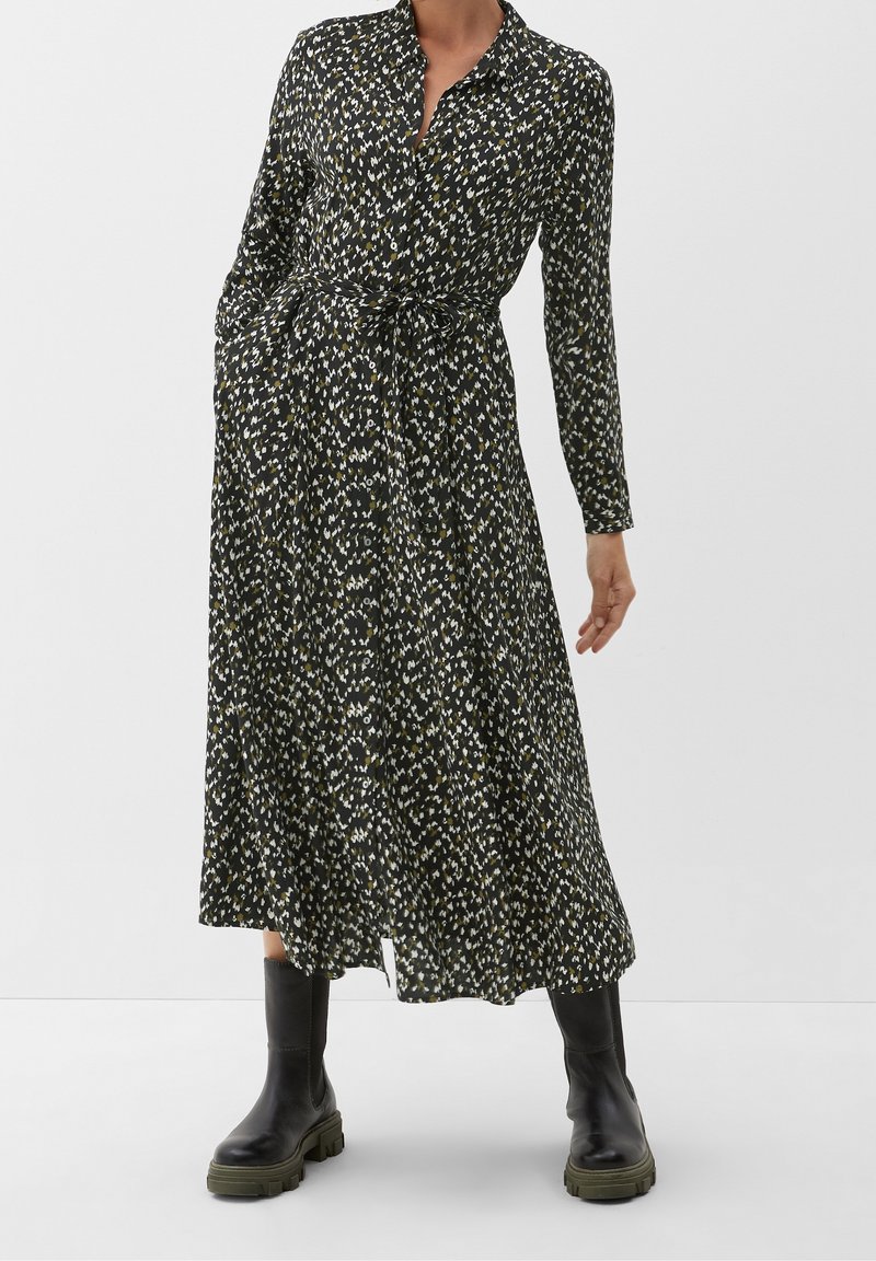 Femme portant une longue robe noire à motifs avec une ceinture à la taille et des bottes noires épaisses, debout devant un fond clair uni.
