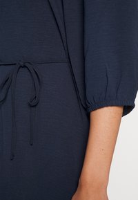 Gros plan d'une personne portant une robe bleu marine avec une ceinture nouée à la taille et des poignets élastiqués aux manches trois-quarts.
