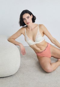 Nizka bralette v svetlo bež in koralni čipki, oboje z nežnimi vzorci. Model sedi na svetli teksturirani površini.