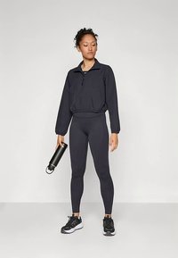 Črn športni pulover s polovičnim zadrgo, elastičnim pasom in dolgimi rokavi, v kombinaciji s črnimi legicami in črnimi supergami, drži steklenico vode.