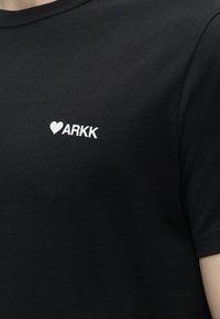 Μαύρο μπλουζάκι με μικρή λευκή καρδιά και το κείμενο "ARKK" τυπωμένο στο αριστερό στήθος, φορεμένο από άτομο με ορατό λαιμό και χέρι.