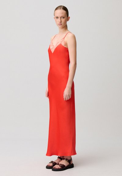 Donna in piedi che indossa un lungo vestito rosso con pizzo ai bordi e sandali neri, sfondo semplice grigio chiaro.