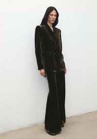 Tmavo hnedý zamatový dvojradový blejzer so ostrými ramenami, v kombinácii s nohavicami s rozšíreným strihom. Minimalistický dizajn a hladká textúra.