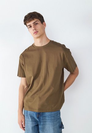 Joven con una camiseta marrón de manga corta y vaqueros azules, de pie frente a un fondo blanco liso con una mano detrás de la espalda.