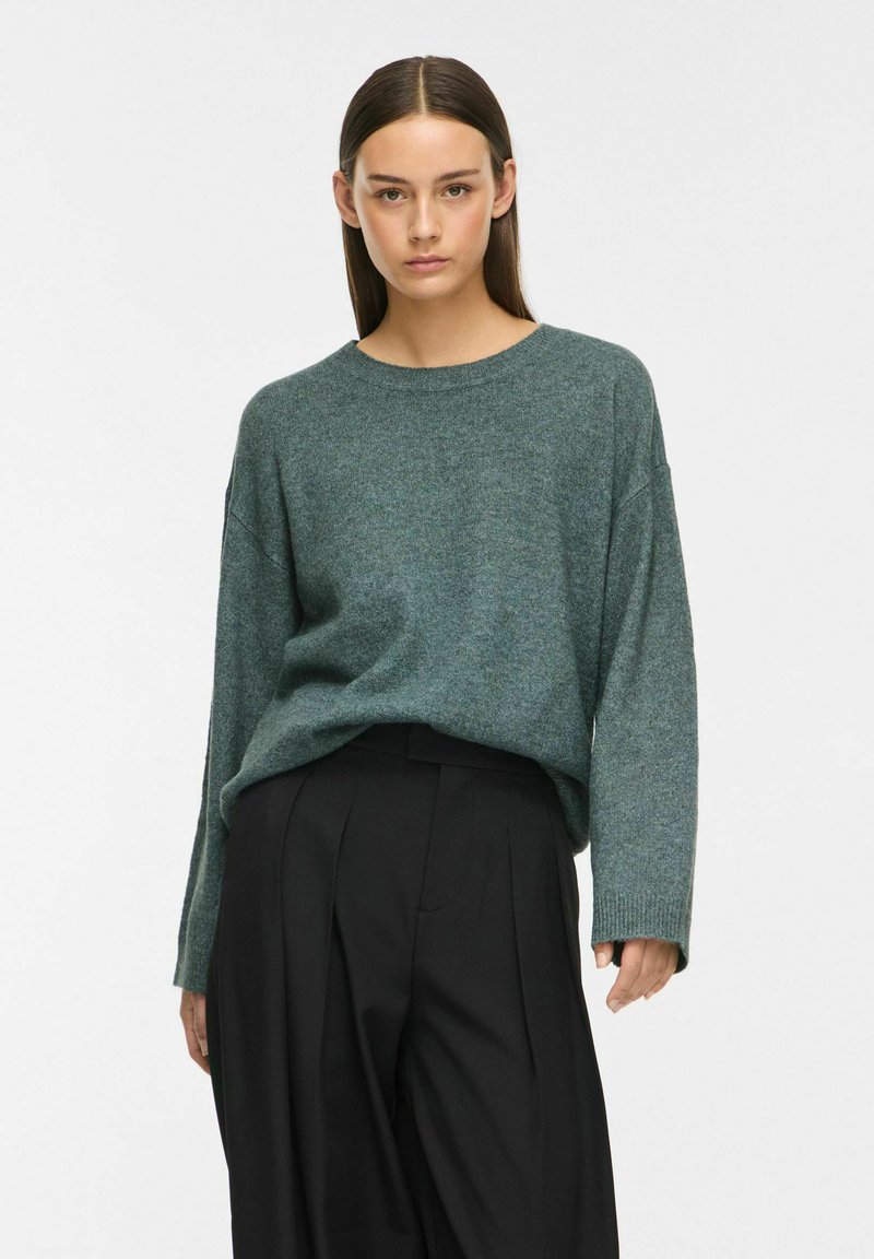 Femme aux cheveux raides et foncés, portant un pull vert ample et un pantalon noir taille haute sur un fond clair uni.