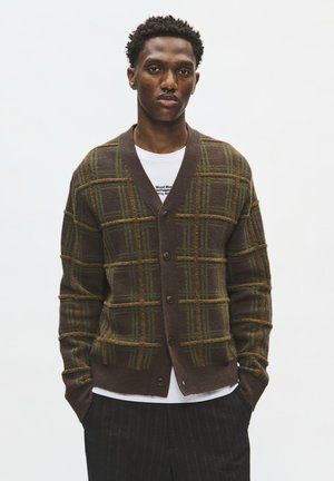 Homme debout, les mains dans les poches, portant un cardigan à carreaux marron sur une chemise blanche et un pantalon sombre à fines rayures, sur un fond uni.