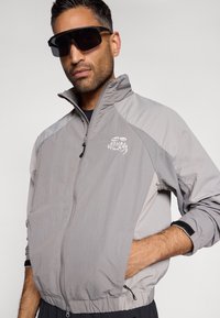 Gaišs pelēks vējjakas mākonis ar augstu apkakli un melniem akcentiem. Iekļauj izšūtu logo uz krūškurvja un rāvējslēdzēja kabatas.