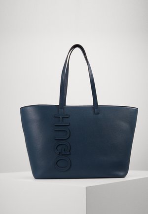 Borsa a mano in pelle blu navy con due spallacci e logo verticale "HUGO" in rilievo sul davanti, posizionata su una superficie bianca da esposizione.