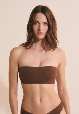 Junge Frau mit heller Haut und braunen Haaren trägt ein trägerloses braunes Bandeau-Top und passenden Bikinihöschen vor einem schlichten Hintergrund.