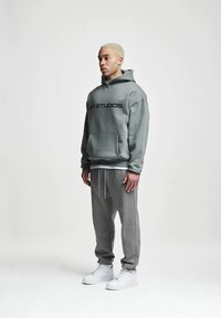 Felpa grigia con cappuccio e tasca frontale, decorata con scritte nere, abbinata a pantaloni della tuta coordinati e sneaker bianche. Tessuto morbido, vestibilità rilassata.