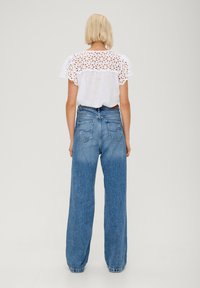 Blusa bianca con dettagli in pizzo a occhio alle spalle, abbinata a jeans azzurri chiari a vita alta e gamba larga. Tessuto morbido, stile casual.