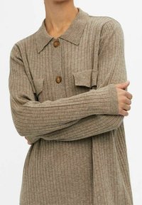 Personne portant un cardigan en tricot côtelé beige avec un col, deux boutons marron et des poches à rabat, les bras croisés sur la poitrine.