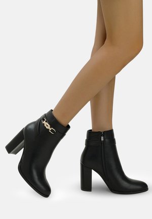 Botas negras hasta el tobillo hechas de cuero sintético, con un detalle de cadena dorada y cierre de cremallera, con un tacón alto grueso y textura suave.