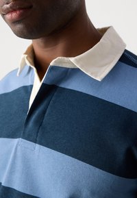 Polo rayé bleu et marine avec un col crème ; tissu doux ; patte de boutonnage ; présente des rayures horizontales contrastées.