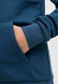 Felpa blu in cotone con tasca a marsupio e polsini a coste. Il tessuto ha una consistenza morbida, con dettagli di cucitura visibili.