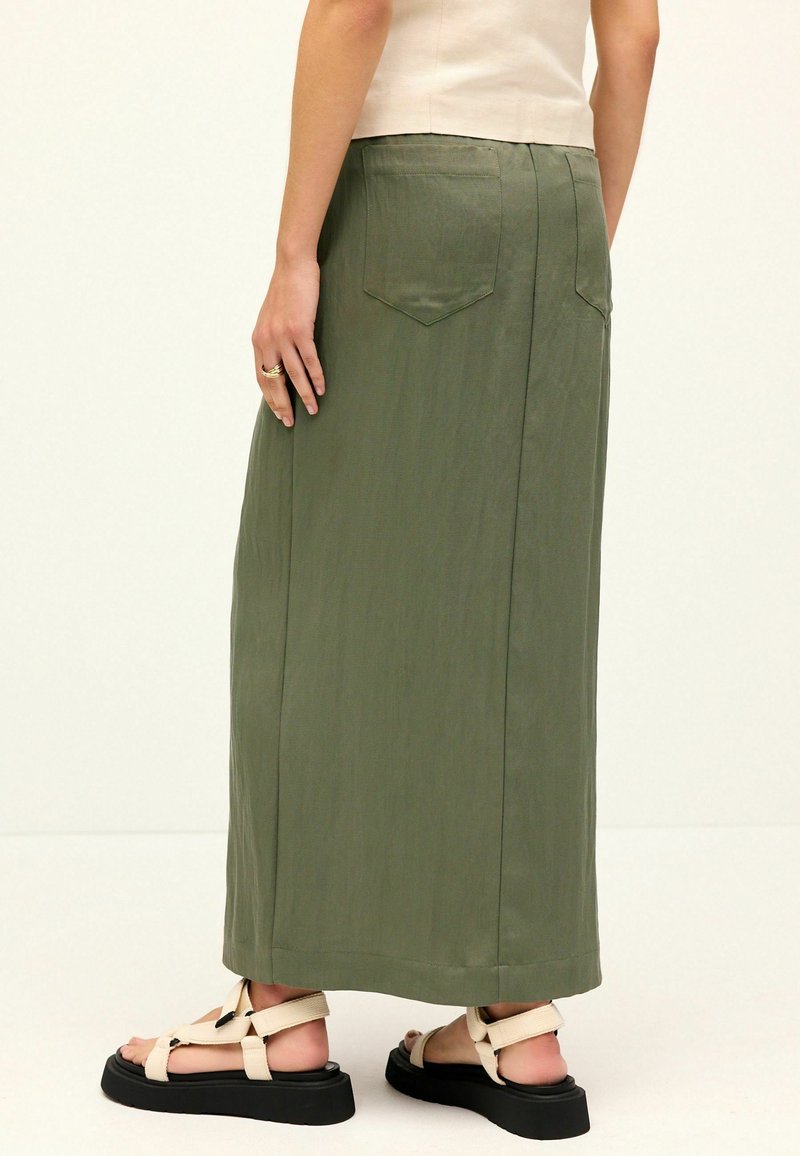 Jupe longue olive, avec une texture lisse, poches arrière et un détail de couture subtil, associée à des sandales à plateforme noires.