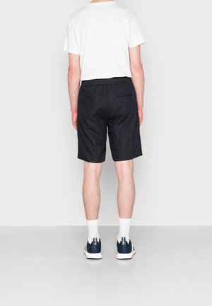 Person wearing black knee-length shorts, white crew socks, navy sneakers, and a plain white short-sleeve T-shirt, standing against a light background.