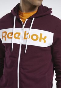 Felpa con zip color marrone bordeaux, con pannello bianco che presenta il logo "Reebok" arancione. Materiale in misto cotone, con cordino regolabile e tasche laterali.
