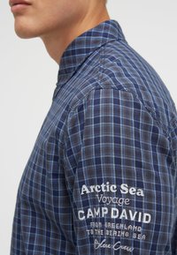 Blau- und grau kariertes Hemd mit hohem Kragen, das mit aufgesticktem Text auf dem Ärmel versehen ist. Leichter Stoff mit glatter Textur.