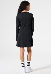 Schwarzes Langarmkleid mit tailliertem Oberteil und ausgestellt Rock, von hinten gezeigt. Kombiniert mit weißen Sneakers und gestreiften Socken.