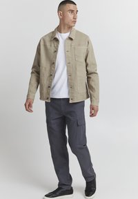 Beige denim jacket with two front pockets, worn over a white t-shirt, paired with dark cargo pants and black sneakers.