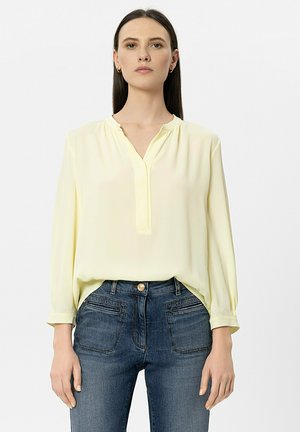 Frau mit langen dunklen Haaren, die eine hellgelbe Bluse mit langen Ärmeln und hoch taillierte blaue Jeans trägt, steht vor einem schlichten Hintergrund.