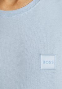 Hellblaues T-Shirt mit Rundhalsausschnitt aus glattem Stoff, mit einem kleinen, in passender Farbe aufgestickten quadratischen Logo auf der Brust.