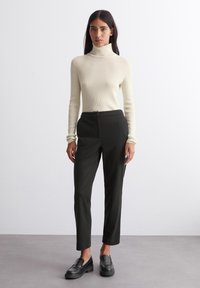 Cremfarbener gerippter Rollkragenpullover kombiniert mit dunkelgrünen, maßgeschneiderten Hosen, die über einen vorderen Knopfverschluss verfügen, und eleganten schwarzen Slippern.