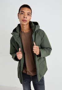 Grønn polstret jakke med hette, med glidelås foran, sidelommer og elastiske mansjetter, båret over en brun skjorte og mørke jeans.