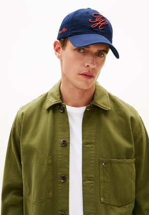 Young man wearing a navy blue cap with red initials and an olive green button-up jacket over a white shirt, facing forward with a neutral expression.