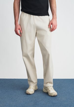 Pantaloni bej din bumbac, cu croială lejeră, având o talie elastică. Purtați cu pantofi sport bej. Detaliu cu suprafață texturată vizibil.