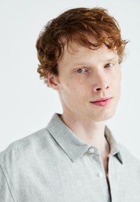 Svetle šedá lněná košile s límečkem, knoflíkovým zapínáním a volným střihem. Materiál má jemnou texturu a subtilní lesk.