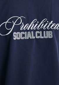Tamsiai mėlynos spalvos medvilninė marškinėliai su išsiuvinėtu balto rašto žodžiu "Draudžiama" ir blokiniais rašmenimis "SOCIAL CLUB" su tekstūrinio paviršiaus apdaila.