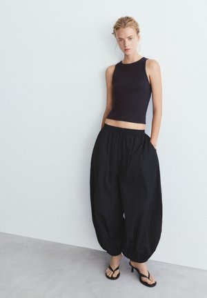 Kobieta w czarnym, bezrękawnym topie, luźnych czarnych spodniach typu balloon i czarnych sandałach na obcasie, stojąca przy białej ścianie.