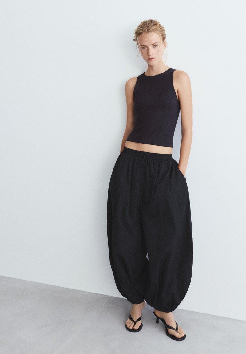 Femme portant un crop top noir sans manches, un pantalon ballon noir large et des sandales à talons noires, debout contre un mur blanc.