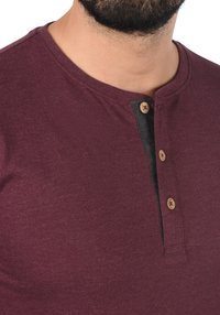 Bordeaux Henley-shirt met drie bruine houten knopen, contrasterende zwarte pat, zachte textuur en een ronde halslijn.